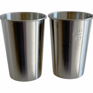 May include: Two stainless steel measuring cups. One cup has "1/2 pt" etched on the side. The cups are cylindrical with a slightly flared top. The metal has a brushed finish.