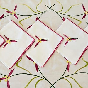May include: Set of six white linen napkins with a pink embroidered floral design. The napkins have a pink border and are folded in a square shape.