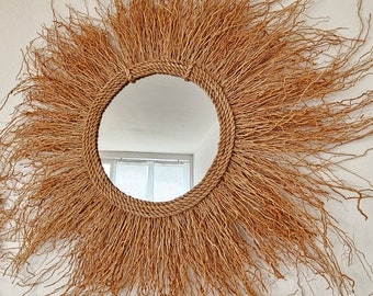 May include: A round mirror with a natural woven frame. The frame is made of thin, light brown strands that create a sunburst effect. The mirror is reflecting a window.