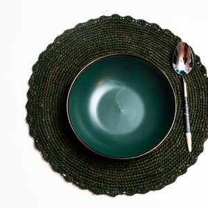 May include: A green ceramic bowl sits on a green woven placemat with a silver spoon beside it.