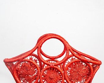 May include: A bright red woven tote bag with a circular handle and a starburst design. The bag is made of thick, woven rope and has a unique, geometric pattern.