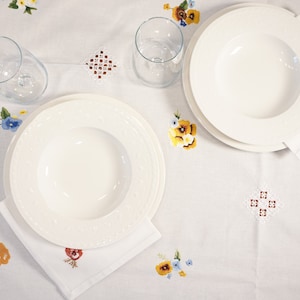 Peut inclure: Deux assiettes plates blanches avec un bord décoratif, l'une sur l'autre, sur une nappe blanche avec des motifs floraux brodés. Un verre se trouve sur la table à côté des assiettes.