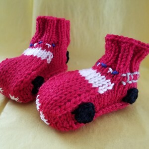 May include: A pair of red knitted baby booties shaped like cars. The booties have white stripes and black wheels.