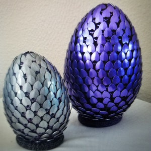 May include: Two decorative dragon eggs. One egg is silver, the other is purple. Both eggs are covered in overlapping, scale-like pieces. The eggs sit on gray, circular bases. The background is a plain, off-white color.