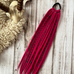 Op de afbeelding: Een bundel fel fuchsia dreadlocks, vastgezet met een zwarte elastiek. De dreads zijn versierd met donkere draadaccenten. De achtergrond toont een rustiek houten oppervlak en schapenvacht.