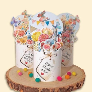 Puede incluir: Tres cajas de regalo cilíndricas blancas con flores de papel y mariposas de colores sobre una rodaja de madera. Cada caja tiene una etiqueta con texto en alemán. Las cajas están decoradas con lunares y atadas con un lazo.