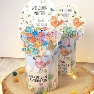 Op de afbeelding: Een pop-upkaart met kleurrijke vogels en de Duitse tekst "WIR ZIEHEN WEITER! DANKE FÜR ALLES" en "WELTBESTE ERZIEHERIN". De kaart toont verschillende vogels in tinten blauw, oranje, groen en roze. De kaart ligt op een houten oppervlak.
