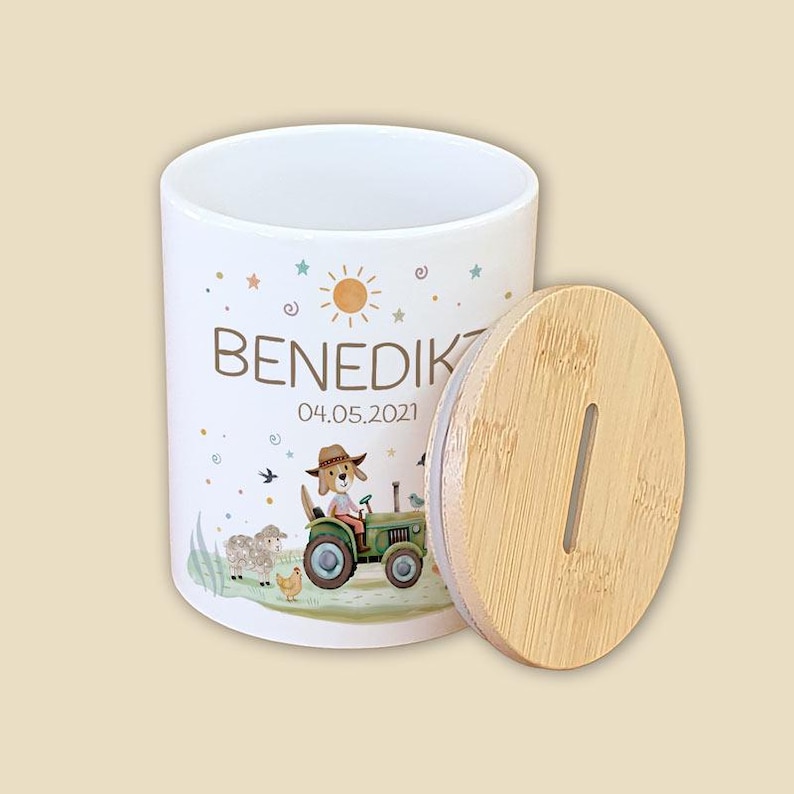 May include: A white ceramic container with a bamboo lid. The container features a cartoon illustration of a dog driving a green tractor, with the name "Benedikt" and the date "04.05.2021" printed on it. The lid has a vertical slot.