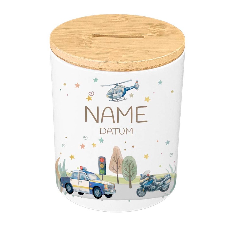 May include: White ceramic money bank with a bamboo lid. The bank features a police car, motorcycle, helicopter, and the words "NAME" and "DATUM". Colorful stars and trees decorate the bank's exterior.