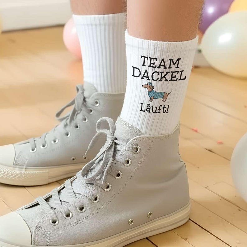 May include: White socks with the text "TEAM DACKEL" and a cartoon dachshund wearing a blue and white striped sweater. The word "Lauft!" is printed below the dachshund. The socks are worn with gray high-top sneakers.