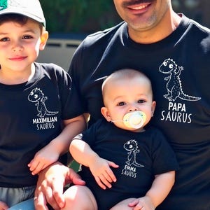 Könnte beinhalten: Drei schwarze T-Shirts mit einem weißen Dinosaurier-Motiv und Text. Die Shirts sagen "Papa Saurus", "Maximilian Saurus" und "Emma Saurus".