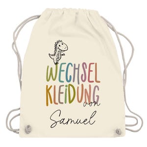 May include: A cream-colored drawstring backpack with a cartoon dinosaur illustration and the text "WECHSEL KLEIDUNG von Samuel" in colorful letters. The bag has gray drawstrings.