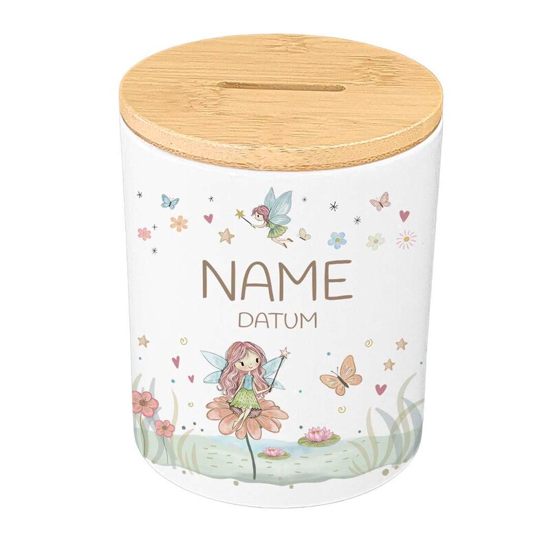 May include: White ceramic money bank with a wooden lid. The bank is decorated with a fairy and floral design, with the words "NAME" and "DATUM" printed on the front. The lid has a slot for inserting coins.
