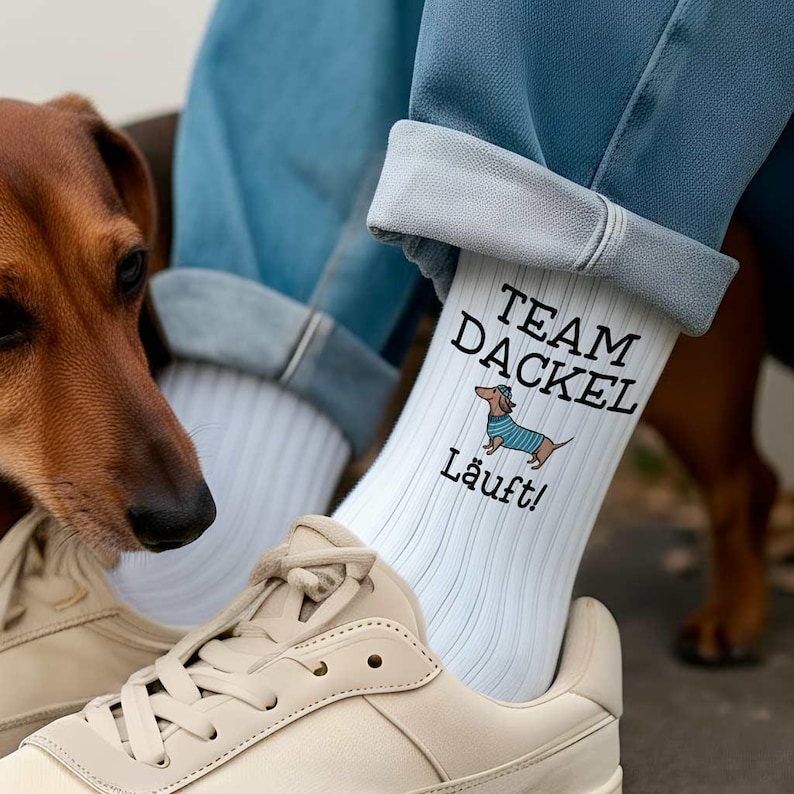 Eine Person trägt weiße Retro-Tennissocken mit einem Dackel-Motiv und dem Spruch 'Team Dackel läuft'. Ein Dackel schaut sich die coolen Socken genauer an.