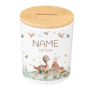 May include: White ceramic money bank with a wooden lid. The bank features a dinosaur theme with illustrations of dinosaurs, a volcano, and the words "NAME" and "DATUM". The design includes green, orange, and blue accents.