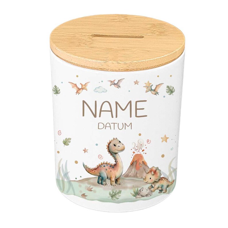May include: White ceramic money bank with a wooden lid. The bank features a dinosaur theme with illustrations of dinosaurs, a volcano, and the words "NAME" and "DATUM". The design includes green, orange, and blue accents.
