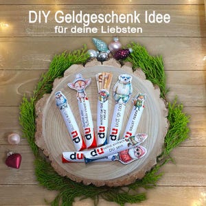 May include: A festive arrangement featuring decorated candy bars with animal-themed toppers, arranged on a wooden slice with greenery and ornaments. The text "DIY Geldgeschenk Idee" is visible at the top.