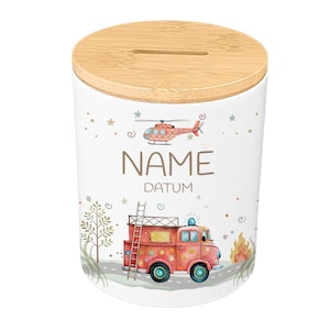 May include: White ceramic money bank with a wooden lid. The bank features illustrations of a red fire truck, a helicopter, and the words "NAME" and "DATUM". The design includes stars, a tree, and flames.
