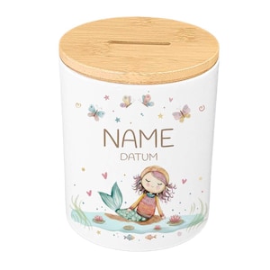 May include: White ceramic money bank with a bamboo lid. The bank features a mermaid illustration, butterflies, and the words "NAME" and "DATUM". The design includes pink, blue, and yellow accents, with a slot for inserting money.