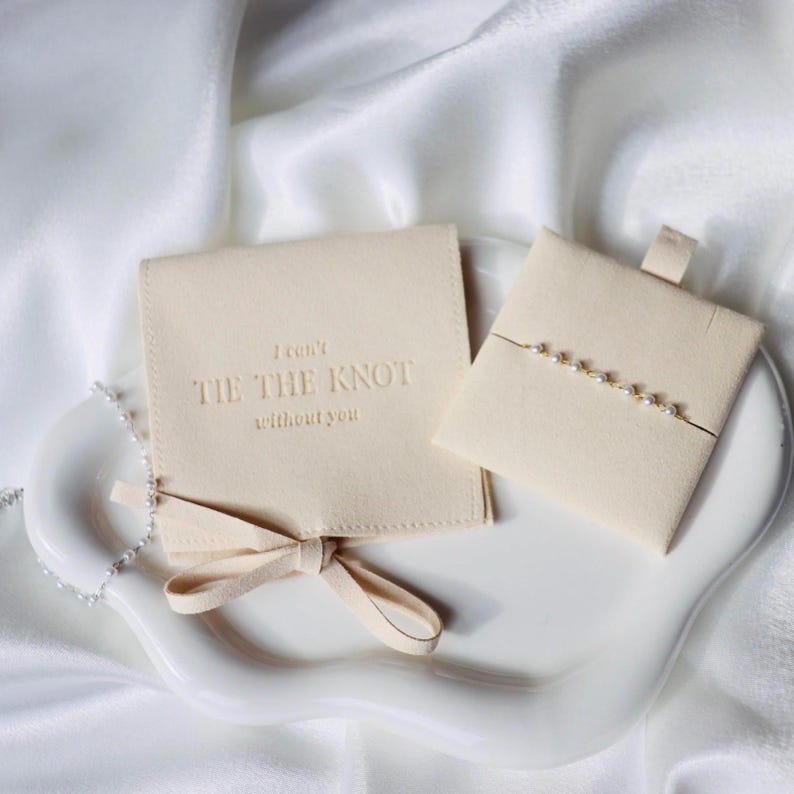 Puede incluir: Una caja de joyer&iacute;a color crema y un peque&ntilde;o tarjetero cuadrado con una pulsera de cadena dorada con perlas. La caja de joyer&iacute;a tiene el texto "I can't TIE THE KNOT without you". Los art&iacute;culos est&aacute;n sobre un plato de cer&aacute;mica blanco.