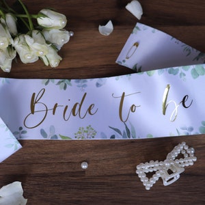 Puede incluir: Banda de satén blanco con un patrón floral verde y letras doradas que dicen "Bride to Be".