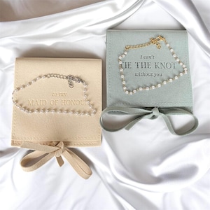 May include: Two square jewellery pouches with ribbon ties, one beige and one sage green. The beige pouch features the text 'to my MAID OF HONOUR' and the green pouch features 'I can't TIE THE KNOT without you'. Each holds a delicate pearl bracelet.