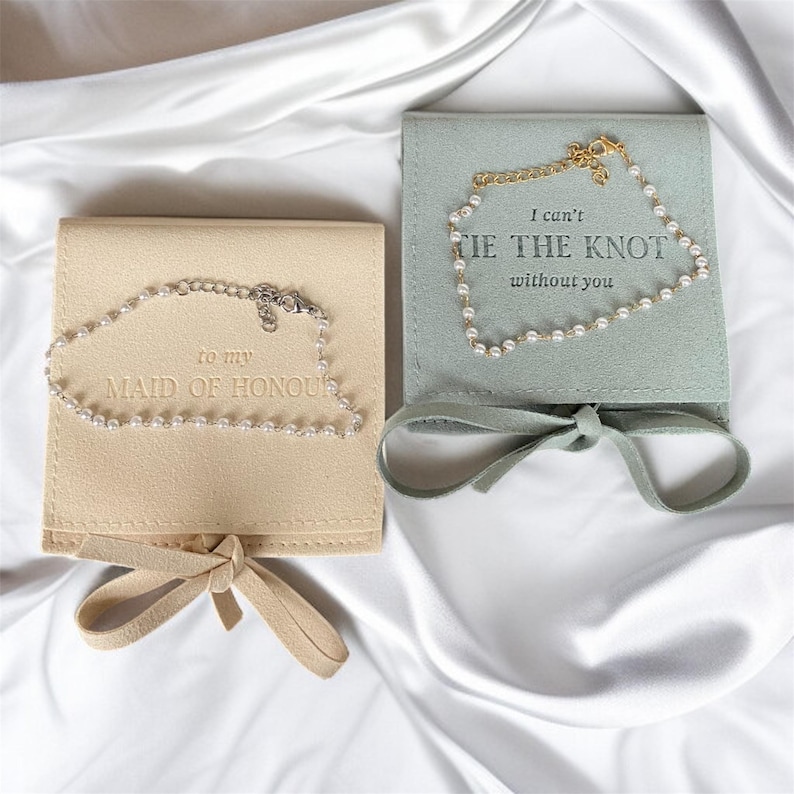 Puede incluir: Dos pulseras de perlas sobre estuches cuadrados. Una es beige y dice "to my MAID OF HONOUR". La otra es verde salvia y dice "I can't TIE THE KNOT without you". Ambos estuches tienen lazos a juego.