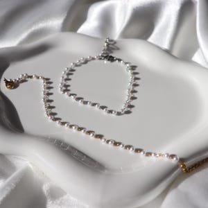 Puede incluir: Una bandeja de cer&aacute;mica blanca para joyas contiene un collar y una pulsera de perlas. El collar tiene cierres dorados y una cadena delicada. Las joyas est&aacute;n sobre una bandeja blanca con bordes ondulados sobre un fondo de tela blanca.