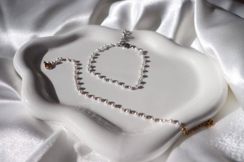 Puede incluir: Una bandeja de cer&aacute;mica blanca para joyas contiene un collar y una pulsera de perlas. El collar tiene cierres dorados y una cadena delicada. Las joyas est&aacute;n sobre una bandeja blanca con bordes ondulados sobre un fondo de tela blanca.