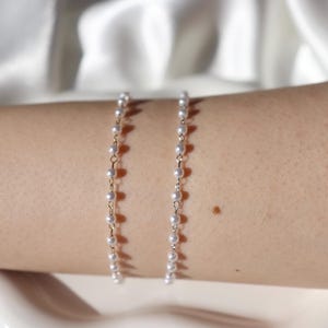 Puede incluir: Se muestran dos delicadas pulseras de perlas con eslabones de cadena dorados. Las pulseras est&aacute;n compuestas por peque&ntilde;as perlas blancas redondas. Las pulseras se muestran en el brazo de una persona, sobre un fondo blanco suave.