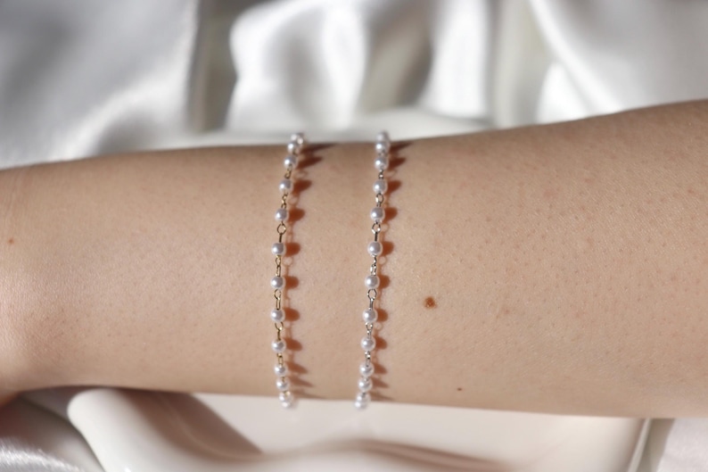 Puede incluir: Se muestran dos delicadas pulseras de perlas con eslabones de cadena dorados. Las pulseras est&aacute;n compuestas por peque&ntilde;as perlas blancas redondas. Las pulseras se muestran en el brazo de una persona, sobre un fondo blanco suave.