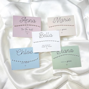 May include: Five rectangular cards in pastel pink, cream, white, light blue, and sage green, each featuring a delicate silver beaded bracelet. The cards display names including Anna, Marie, Bella, Chloe, and Diana, along with small heart icons.