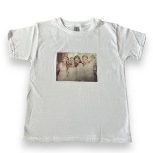 May include: White t-shirt with a printed image of four women in white dresses standing in a room.