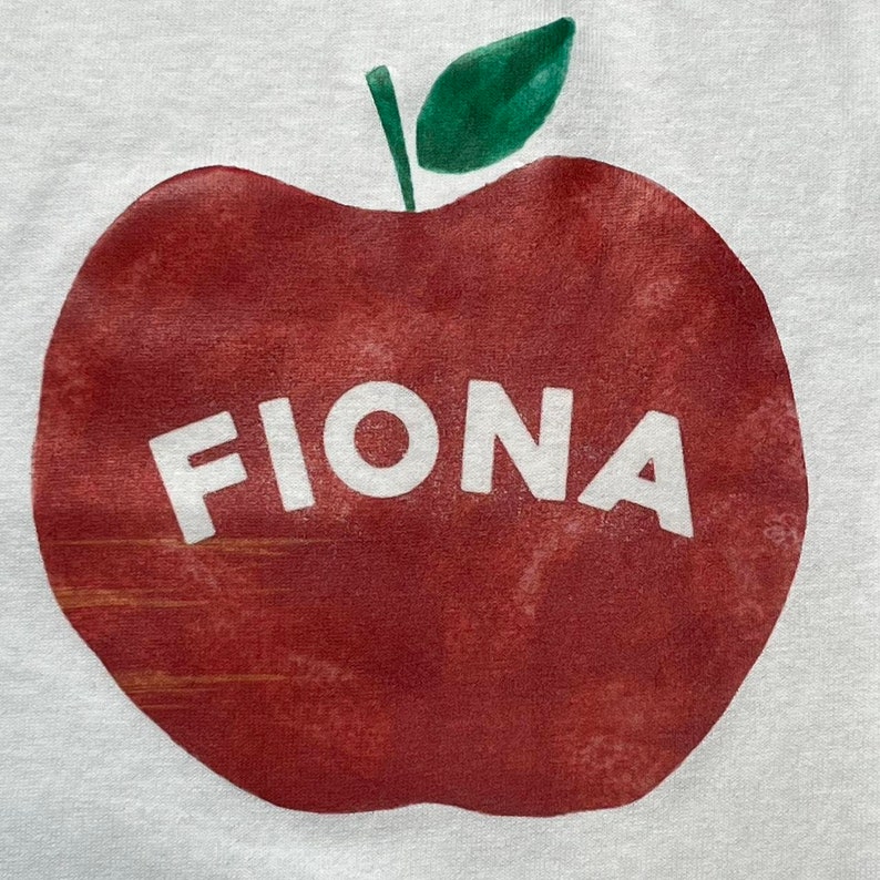 May include: A red apple with a green leaf on top. The word "FIONA" is printed in white on the apple.