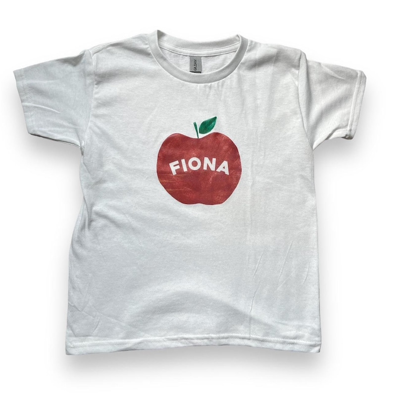 May include: White t-shirt with a red apple graphic and the name "Fiona" printed on it.