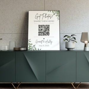 May include: A white sign with green foliage on the sides. The sign says "Got Photos? Share them here!" with a QR code and the text "Garrett & Ashley July 21, 2023".