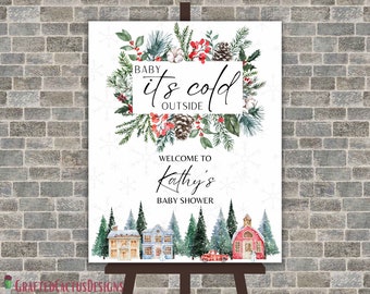 May include: A white baby shower welcome sign with a winter theme. The sign features a watercolor illustration of a snowy village with houses, trees, and a red truck. The text on the sign reads "Baby it's cold outside" and "Welcome to Kathy's Baby Shower".