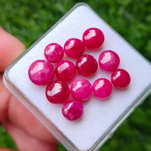 Op de afbeelding: Een verzameling van elf kleine, ronde, gepolijste cabochon edelstenen in een levendige, dieproze-rode kleur. Deze gladde, koepelvormige stenen zijn gerangschikt op een witte schuimrubberen inzet in een vierkant, doorzichtig plastic doosje.