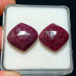 Könnte beinhalten: Zwei Rubine im Kissenschliff mit tiefroter Farbe und facettierten Oberflächen. Die Edelsteine sind nebeneinander in einem durchsichtigen Kunststoffbehälter mit einer weißen Schaumstoffeinlage zur sicheren Aufbewahrung platziert.