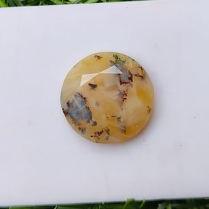 May include: A round, faceted gemstone with a translucent, pale yellow and amber base color. The stone features dark, dendritic, fern-like inclusions that branch across its surface, creating a natural, intricate pattern within the polished material.