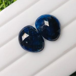 Könnte beinhalten: Zwei oval geformte, tiefblaue Edelsteine mit facettierter Oberfläche. Jeder Stein hat ein poliertes Finish, das das Licht reflektiert und eine dunkle, gesättigte blaue Farbe über die gesamte geschliffene Oberfläche der Steine hinweg zeigt.
