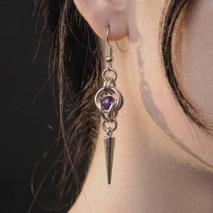 May include: A silver earring with a purple bead and a cone-shaped dangle. The earring is made of multiple linked rings and is shown against a dark background.