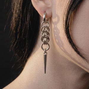 Könnte beinhalten: Silberner Ohrring mit einer Reihe von verbundenen Ringen und einem spitz zulaufenden Kegelanhänger. Der Ohrring wird im Ohr getragen, wobei sich das Design nach unten erstreckt. Das Metall hat eine polierte Oberfläche, die Licht reflektiert.
