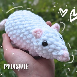 Può includere: Un topo di peluche bianco fatto a mano con orecchie e naso rosa e occhi neri. Il peluche è tenuto in mano, con la scritta "RAT PLUSHIE" in basso. Lo sfondo è verde sfocato.