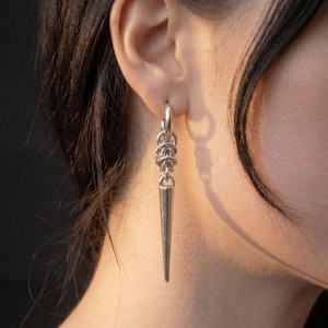 Könnte beinhalten: Ein silberfarbener Ohrring mit einem glatten, spitz zulaufenden Anhänger, der an einem kleinen runden Creolen-Ring hängt. Der Dorn ist durch eine Reihe ineinandergreifender Metallringe, die ein kurzes, strukturiertes Kettenglied bilden, mit dem Ring verbunden.