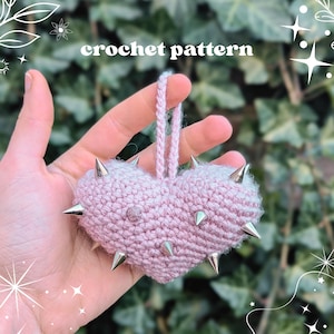 May include: A crocheted pink heart with silver spikes. The heart-shaped ornament has a loop for hanging and the words "crochet pattern" in white text above it. The background is a blurred green.