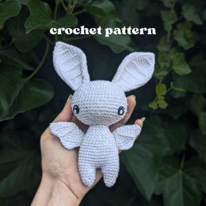 Könnte beinhalten: Ein weißes gehäkeltes Fledermaus-Spielzeug mit großen spitzen Ohren, kleinen Flügeln und schwarzen Sicherheitsaugen. Der Text "crochet pattern" ist über dem Spielzeug zu sehen. Die Fledermaus hat eine strukturierte Garnoberfläche und einen runden Kopf.