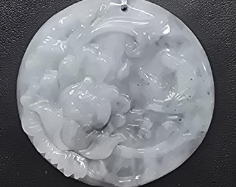 Puede incluir: Colgante redondo de jade verde pálido con detalles tallados de flores y figuras. Cuelga de un cordón negro con detalles metálicos plateados y una cuenta roja. El fondo es gris oscuro.