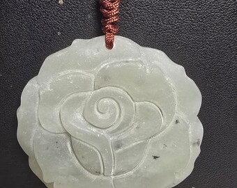 Puede incluir: Un colgante de jade tallado en forma de rosa. El colgante es de color verde claro y tiene un cordón marrón atado a él.