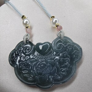 Puede incluir: Un collar con un colgante de jade tallado con un dise&ntilde;o floral y un coraz&oacute;n en el centro. El colgante est&aacute; unido a un cord&oacute;n azul claro con cuentas blancas y rosadas.